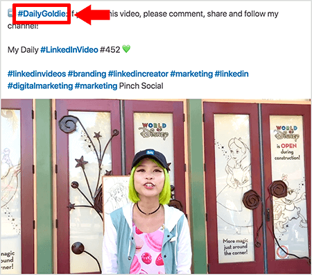 Šis ir ekrānuzņēmums, kas ilustrē to, kā Goldija Čana izmanto hashtagus savu LinkedIn video ierakstu tekstā. Sarkanās norādes tekstā norāda uz #DailyGoldie hashtag, kas ir unikāls tikai viņas video ierakstiem un palīdz izsekot kopīgošanu. Ziņā ir arī citas atbilstošas ​​atsauces, kas palīdz cilvēkiem atrast viņas videoklipu, tostarp #LinkedInVideo. Video attēlā Goldija stāv pie dažām durvīm Disneja pasaules izstādē. Viņa ir Āzijas sieviete ar zaļiem matiem. Viņai ir melna LinkedIn cepure, melna kakla kaklarota, rozā krekls ar macaron apdruku un zila un balta jaka.